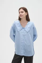 Camisa de algodón color celeste, con cuello grande tipo babero con volados, cierre frontal con botones y mangas largas con puños.