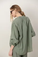 Camisa de algodón verde con bordados al tono, escote en V y cuello camisero corto, mangas tres cuartos abullonadas con puños elásticos y cierre frontal parcial con botones.