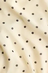 Vestido corto de gasa transparente en color beige con estampado de lunares negros. Presenta un diseño con escote halter, tiras anchas para atar en la nuca, espalda abierta, cintura elástica fruncida y falda escalonada con vuelo. Incluye forro interior.