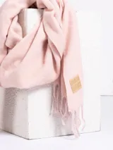 Bufanda de tejido suave en color rosado, con diseño liso y terminaciones con flecos en los extremos. Incluye una etiqueta de cuero sintético con el logo de la marca en uno de los bordes.