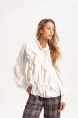 Camisa de gasa color blanco con volados frontales y en las mangas, cuello con lazo y mangas largas abullonadas con puños ajustados.