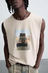 Camiseta tank de punto color beige claro, corte relajado (relaxed fit) y cuello redondo. Presenta un estampado central de un vehículo todoterreno cargado de equipaje conduciendo por una carretera desértica. Los bordes del cuello y las sisas tienen un acabado deshilachado.