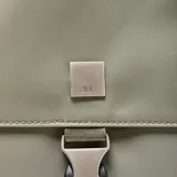 Mochila de diseño minimalista en color verde oliva, confeccionada en material textil suave. Cuenta con un compartimento principal con cierre de doble cremallera, un bolsillo frontal con solapa y cierre de hebilla tipo clip, y un asa superior reforzada.