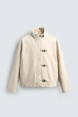 Cazadora de algodón color beige claro, corte relajado, con cuello subido y manga larga. Cierre frontal con cremallera oculta por solapa con cierres metálicos tipo alamar.