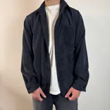 Sobrecamisa de pana negra con cierre frontal de botones, cuello clásico, mangas largas con puños abotonados y un bolsillo en el pecho.