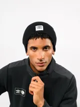 Gorro tipo beanie de tejido de punto acanalado en color verde oliva, con parche frontal rectangular blanco que presenta el logo de Umbro en rojo.
