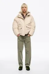 Chaqueta acolchada tipo puffer de corte globo en color beige, con cuello subido, capucha, cierre frontal con cremallera y botones de presión, y bolsillos delanteros con tapeta.