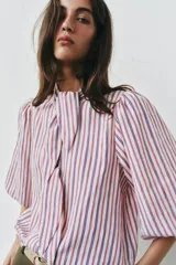 Camisa de rayas verticales rojas, blancas y azules, con cuello alto decorado con lazada, mangas abullonadas por debajo del codo y cierre frontal de botones forrados.