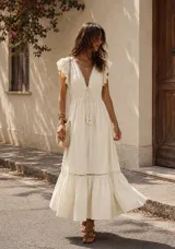 Vestido midi de color beige, con escote en V profundo, mangas cortas con volados y cintura elástica ajustable con cordones y borlas. Presenta detalles de encaje en el escote y en el ruedo, con una falda amplia y fluida.