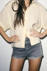 Shorts de jean de algodón, tiro alto, color azul claro desgastado. Presenta cinco bolsillos, cierre frontal con botón metálico y cremallera, y bajo desflecado con vuelta.