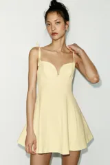 Vestido corto color amarillo pálido, con escote corazón, tirantes finos ajustables con detalle de lazos y falda acampanada.