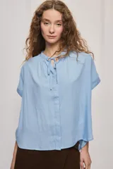 Camisa de manga corta color celeste, confeccionada en viscosa fluida, con silueta amplia y escote redondo con frunces y lazo para anudar.