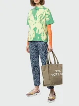 Bolso tipo tote de Marc Jacobs, confeccionado en lona resistente de color negro. Presenta el nombre del modelo estampado en blanco en la parte frontal y el logo de la marca debajo. Cuenta con doble asa superior para llevar al hombro o en la mano.