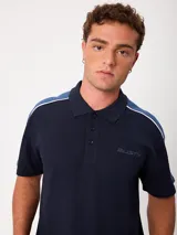 Remera polo gris melange de ajuste holgado, con cuello y cierre frontal de botones. Presenta detalles en azul marino y blanco en los hombros y mangas, y el logo de la marca bordado en el pecho.