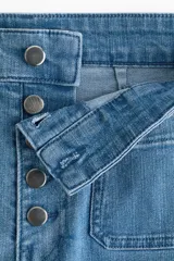 Jeans de tiro alto y corte acampanado, confeccionados en denim con leve elasticidad. Presentan cierre frontal con múltiples botones metálicos visibles, bolsillos de parche delanteros y diseño de perneras anchas desde la rodilla.