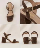 Sandalias de gamuza color marrón chocolate, de taco cuadrado medio y punta cuadrada. Tienen una tira delantera ancha y una tira ajustable al tobillo con hebilla metálica dorada.