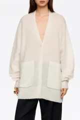 Cardigan largo de corte oversize, color blanco, con escote en pico, cierre frontal de botones y bolsillos de plastrón. Presenta mangas largas con puños acanalados.