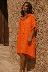 Vestido camisero corto de lino color naranja, de diseño amplio y cómodo, con cuello camisero, cierre frontal con botones de nácar y mangas largas con trabilla para sujetarlas arremangadas. Presenta un corte asimétrico, siendo más largo en la parte trasera.