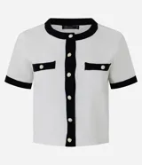 Cardigan de punto de manga corta con cuello redondo y cierre frontal de botones perolados. Presenta un diseño bicolor en blanco y negro con detalles de contraste en cuello, puños, tapetas de bolsillos frontales y vista central.