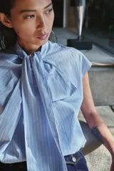 Camisa sin mangas confeccionada en algodón, con cuello redondo y detalle de lazada frontal. Presenta un diseño de corte holgado con bajo redondeado y cierre posterior mediante abertura y botones.