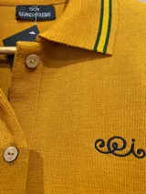 Cárdigan de punto de lana merino color mostaza con cuello tipo polo, cierre frontal de botones y detalles de rayas verdes y amarillas en los puños y el borde inferior. Cuenta con un bordado sutil en el pecho.