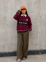 Sweater de punto Nude Project modelo Legend Football Knit Polo, con diseño a rayas en tonos bordó, negro y gris, cuello polo gris claro con cierre frontal parcial, parche bordado de rosa celeste y la inscripción Nude Project en el pecho.