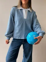 Buzo tipo chomba de corte oversize con diseño a rayas horizontales celestes y blancas. Presenta cuello camisero blanco y un bordado en la espalda con la inscripción República Oriental del Uruguay.