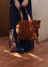 Bolso tipo shopper confeccionado en cuero descarne color marrón, con diseño de flecos en los laterales y correas ajustables con hebillas metálicas. Incluye doble bolsillo interno y tira porta llaves.