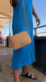Bandolera pequeña de diseño rectangular en color beige, confeccionada en cuero sintético. Presenta una correa larga ajustable del mismo material y cierre superior con cremallera. Incluye el logo de la marca grabado en la parte frontal.