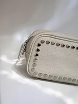 Cartera bandolera de cuero sintético color beige, con diseño rectangular y detalle de tachas metálicas circulares en el contorno frontal. Cuenta con correa larga ajustable y desmontable mediante mosquetones, cierre superior y bolsillos internos y externos.