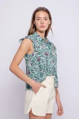 Camisa sin mangas color crudo con estampado floral negro y verde, cuello camisero, cierre frontal con botones y volados en los hombros.