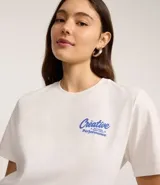 Remera de algodón color blanco tiza, manga corta y cuello redondo. Presenta un estampado grande en la espalda con la palabra "Creative" en tipografía cursiva iridiscente, un oso de goma con efecto holográfico en tonos azules y rosados, y el texto "MATTERS MORE THAN P¡FORMACE" debajo.