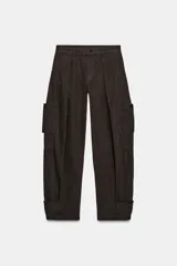 Pantalón cargo de algodón color marrón oscuro, tiro medio, con pliegues frontales, bolsillos delanteros ocultos y bolsillos de plastrón con solapa y botones a presión en las piernas. El bajo del pantalón tiene una trabilla con botón y se muestra doblado hacia afuera.