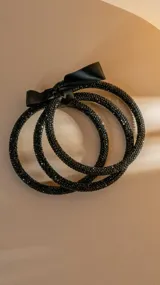 Set de tres pulseras rígidas negras cubiertas de brillos, unidas por un lazo de satén negro.