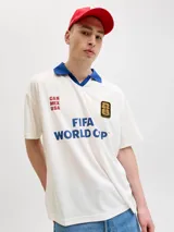 Camiseta de manga corta con cuello tipo polo en color azul, cuerpo en color blanco y calce relajado. Presenta estampados frontales con la leyenda 'FIFA WORLD CUP' y 'CAN MEX USA', junto con un parche conmemorativo del evento.