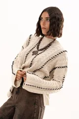 Buzo de tejido de punto color crudo con diseño de trenzas y costuras decorativas en color negro que recorren la prenda en forma de espiral. Presenta cuello redondo y mangas largas con terminación acampanada.