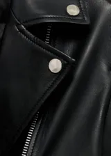 Campera estilo biker de cuero ovino negro, con solapas amplias, cierre frontal asimétrico, bolsillos laterales con cremallera y detalles de tachas metálicas en el cuello.