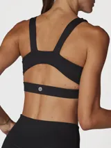 Top deportivo negro con escote redondo, tirantes anchos y un corte debajo del busto que crea una abertura. Incluye logo blanco en el pecho.