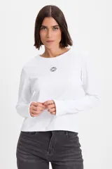 Remera de manga larga ajustada al cuerpo, confeccionada en tejido acanalado blanco. Presenta cuello redondo, aberturas para los pulgares en los puños y un pequeño bordado decorativo en el centro del pecho.