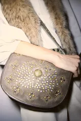 Cartera tipo bandolera de cuero vacuno color gris con diseño redondeado. Presenta una solapa frontal decorada con tachas metálicas en tonos dorados y plateados formando un patrón ornamental, incluyendo un aplique central circular grabado.