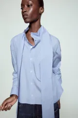 Camisa de algodón color celeste, con cuello alto y lazo integrado en el mismo tejido. Presenta cierre frontal con botones, manga larga con puños y un bolsillo frontal en el pecho. Diseño de corte recto.