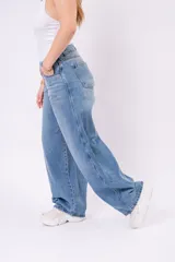 Pantalón de jean wide leg en lavado azul claro vintage, con corte amplio y fluido. Presenta un cierre asimétrico con botones expuestos en la parte delantera, talle alto y bolsillos funcionales.