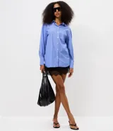 Camisa alargada de manga larga, confeccionada en tricolina de algodón con diseño de rayas finas azules y blancas. Cuenta con cuello inglés, cierre frontal con botones y un bolsillo aplicado en el pecho.