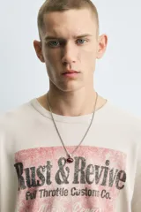 Camiseta de punto de algodón con corte boxy fit, cuello redondo y manga corta. Presenta un estampado frontal con temática de taller mecánico en tonos rojo desgastado y negro, incluyendo el texto "Rust & Revive", "Old Iron Garage" y detalles técnicos. La prenda tiene un efecto lavado y acabados irregulares.