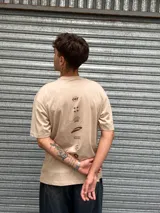 Remera de corte oversize color beige, con cuello redondo y mangas cortas. Presenta un diseño gráfico estampado en el pecho con la palabra 'OSTEN' y logotipos abstractos en color marrón.