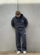 Conjunto de dos piezas compuesto por un buzo con capucha y cierre frontal y un pantalón de jogging, ambos confeccionados en tejido con efecto acid wash en color gris oscuro. El buzo cuenta con bolsillos frontales y logo bordado, mientras que el pantalón presenta un corte holgado y logo bordado en la pierna.