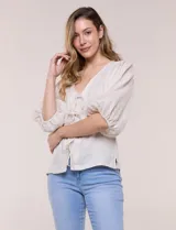 Blusa color beige confeccionada en lino, con escote en V pronunciado y cierre frontal de múltiples lazos para atar. Presenta mangas abullonadas tres cuartos con puños elásticos y corte relajado.