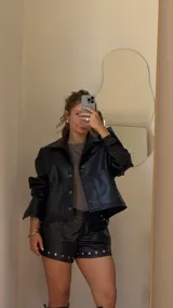 Campera corta negra de ecocuero con cuello camisero, cierre frontal con botones a presión, dos bolsillos con solapa y mangas largas con puños abotonados.