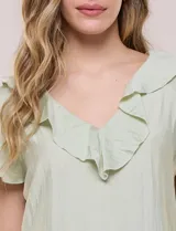 Blusa de satén texturizado color verde menta, con escote en V y volados que bordean el cuello. Presenta mangas cortas acampanadas y doble nivel de volados en la parte inferior, formando una silueta péplum.