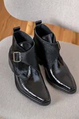 Botin de cuero negro con acabado brillante y detalle de solapa en cuero con textura de pelo, cierre con hebilla metálica lateral y punta fina.
